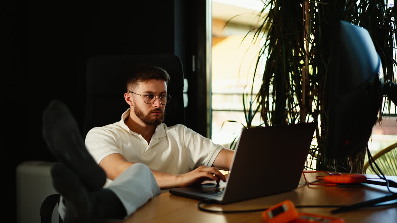 Relaxed young man working on a laptop from home with feet up, in a cozy modern workspace. Concept of freelance lifestyle, remote work, and balanced productivity