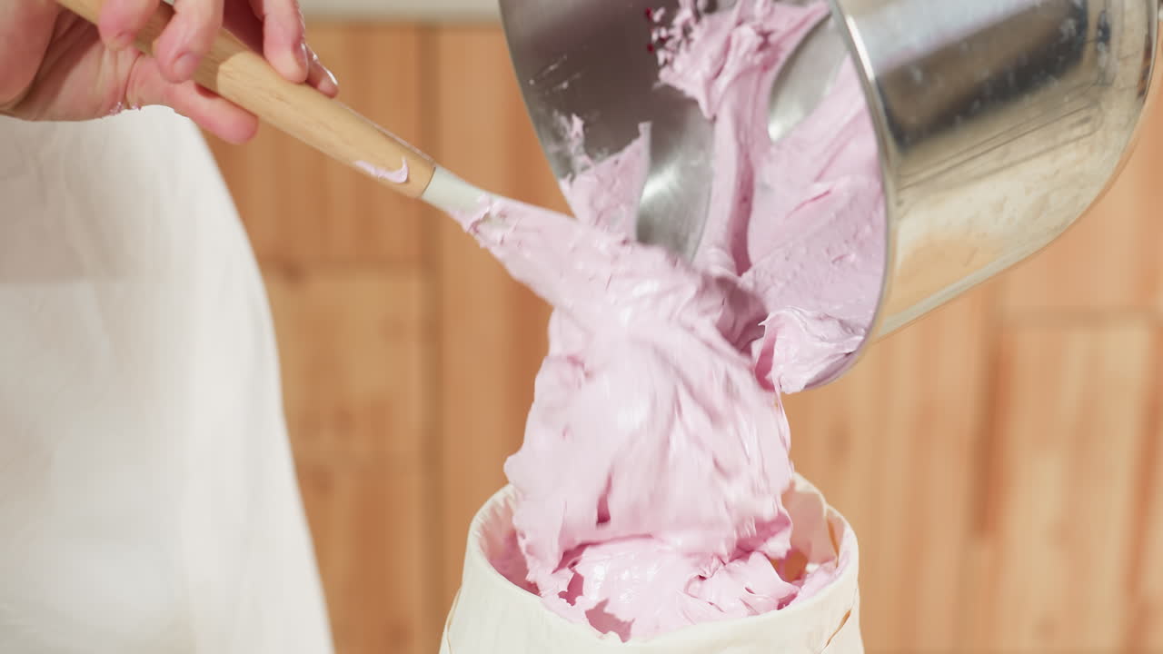 Baker scrapes fluffy pink dough from large mixing bowl using wooden spatula and fills piping bag, preparing for baking in cozy kitchen with rustic decor, soft lighting, and baking setup