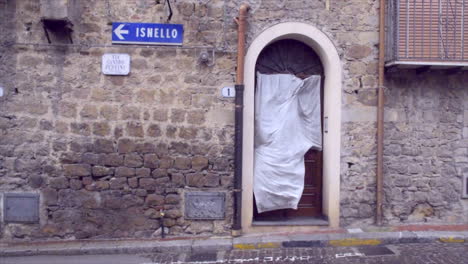ancient, traditional Sicilian house with a weathered facade. The wind blows a curtain on the window. The wall features a street sign pointing to Isnello,
