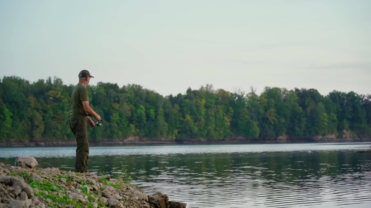 Senior fishing in the river from coast at dusk. Verdant forest in the background.
