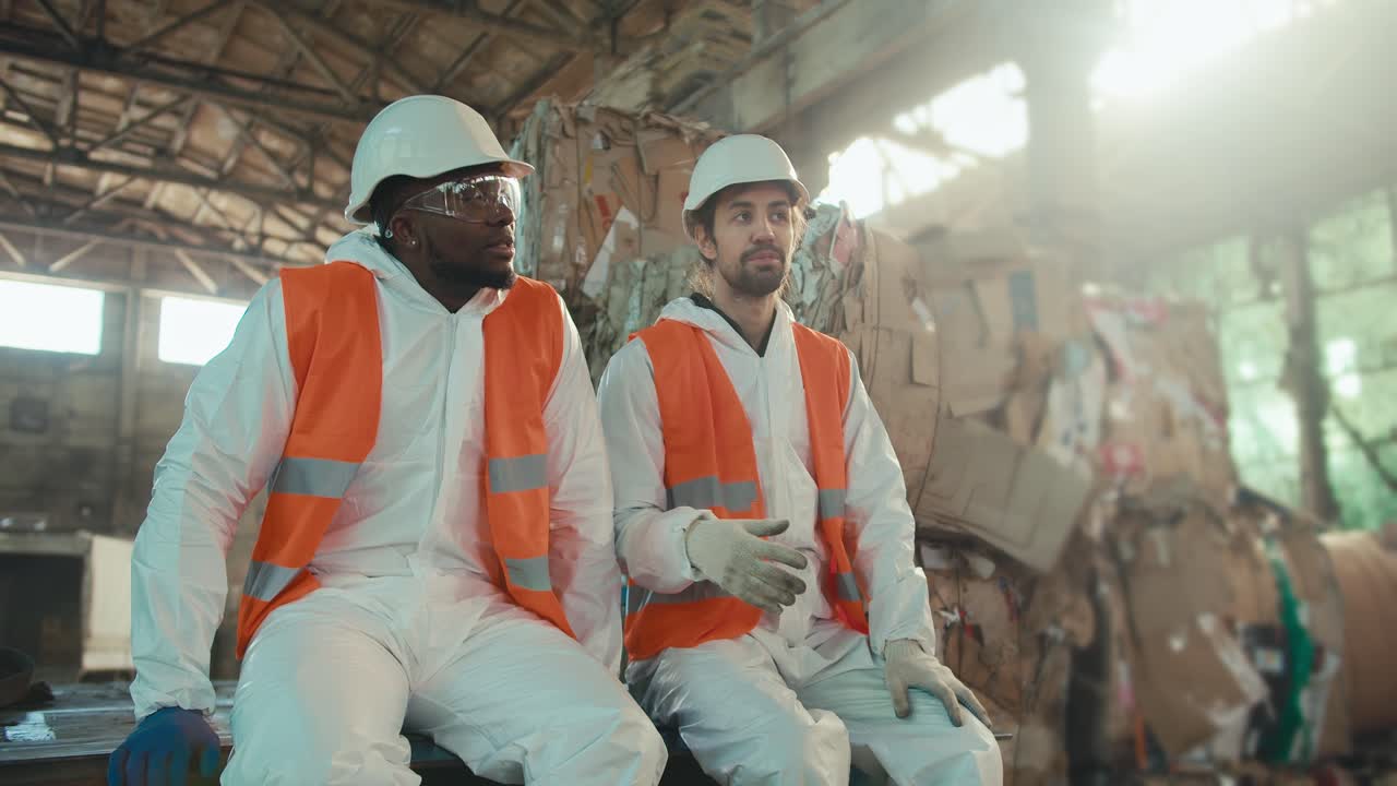 A Black man in a white uniform and an orange vest sits with his brunette male colleague with a beard in a white helmet and discusses during a break at a large waste and paper recycling plant