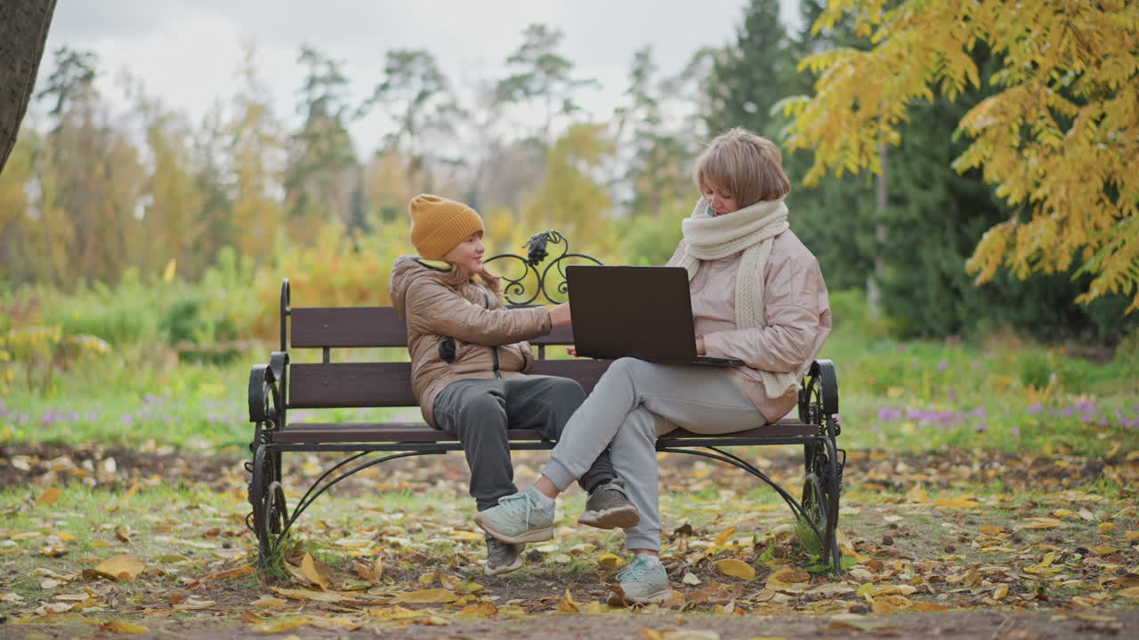 woman sits on bench working on laptop outdoors in autumn park while little girl approaches holding foliage to distract mother and mother gently shifts hand aside to avoid interruption