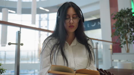 warm glow surrounds young woman with headphones and glasses as she reads book quietly in modern mall interior, seated on artistic bench with glass railing and blurred shoppers in background