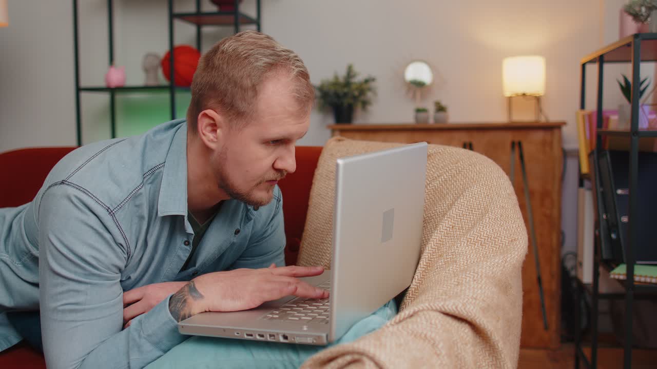 Adult man using laptop computer lying on sofa at home working online shopping watching movies