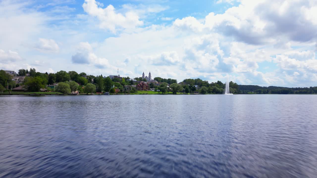 Expansive drone shot looking over a wide Baltic lake with distant town and fountain framed by trees and open skies. Shot in Zarasai, Lithuania