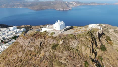 drone view in greece circling around a white church at the top of a brown hill with a greek white house town facing blue sea on a mountain