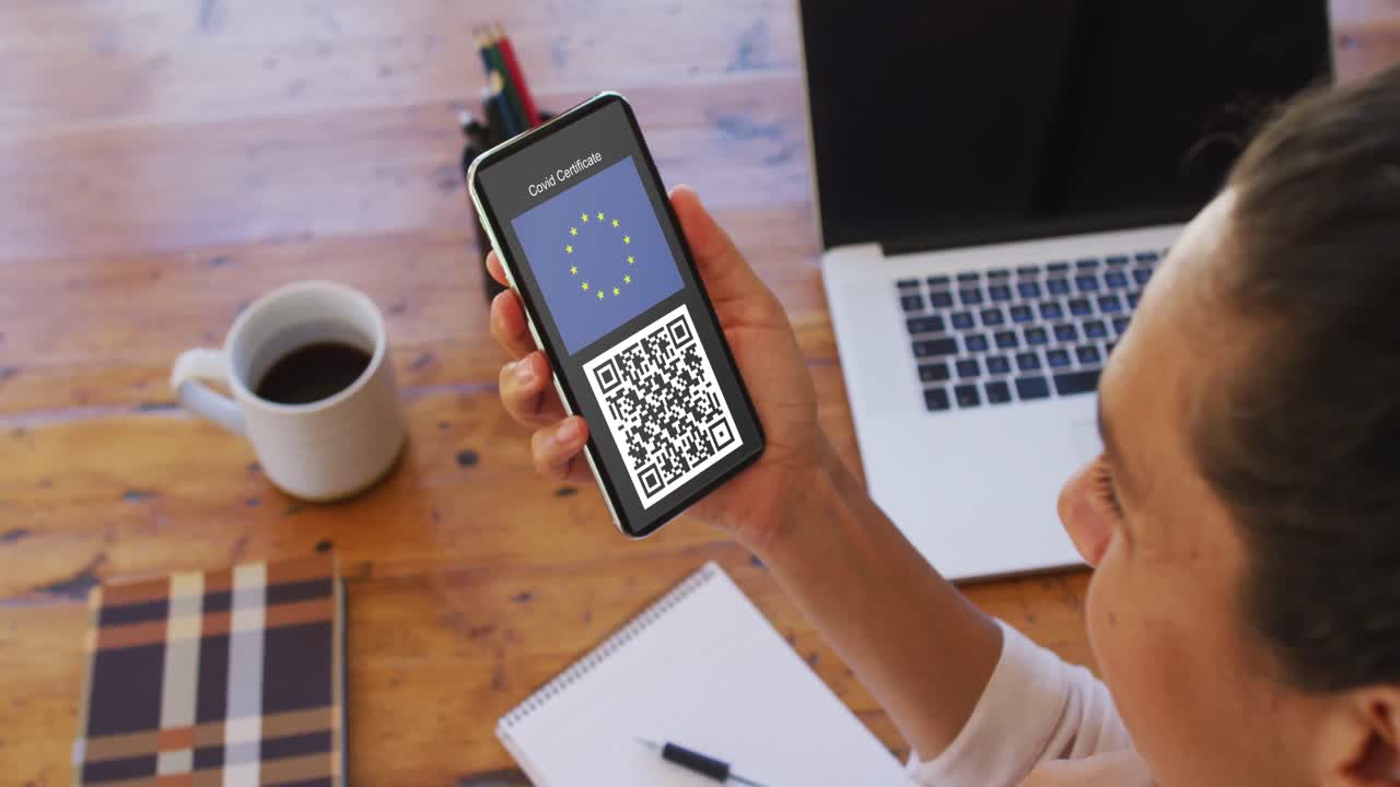 Woman at desk holding smartphone with covid vaccination certificate, eu flag and qr code on screen