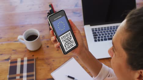 Woman-at-desk-holding-smartphone-with-covid-vaccination-certificate,-eu-flag-and-qr-code-on-screen