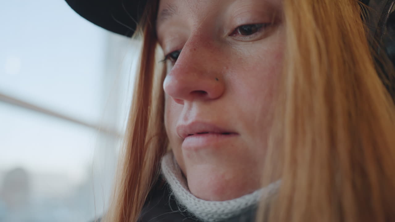 Pensive white woman on train window, redhead wearing hat and headphones, wool scarf pulled up, freckles visible, natural light through glass, closeup profile shot capturing quiet introspection, urban