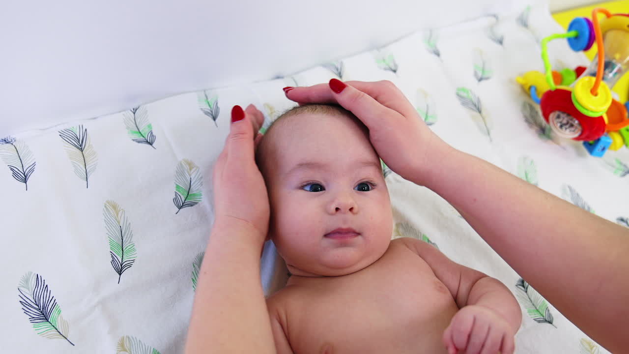 Doctor's hands checking baby's head, touching him gently and carefully. Cute baby is watching the pediatrician intently. Close up.