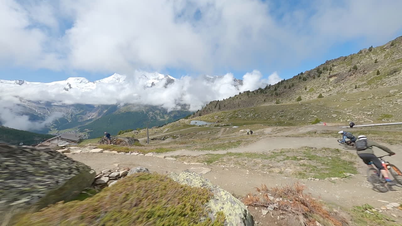 FPV drone follows two mountain bikers descending steep, exposed alpine trails above Saas-Grund and Saas-Fee on a clear bluebird summer day, showcasing epic terrain and speed