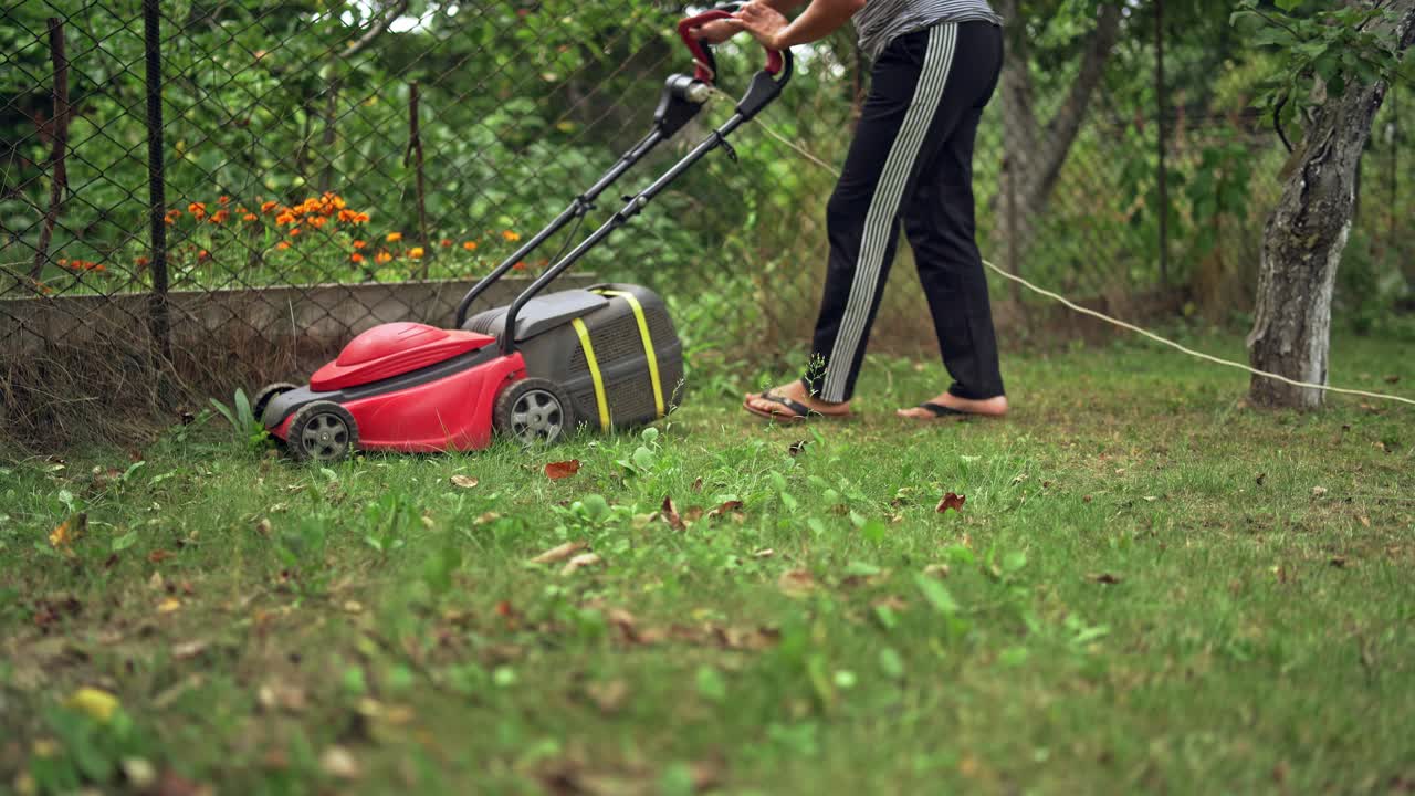 Woman working with electic mower machine in the garden. Lawn mower cutting green grass outdoors.