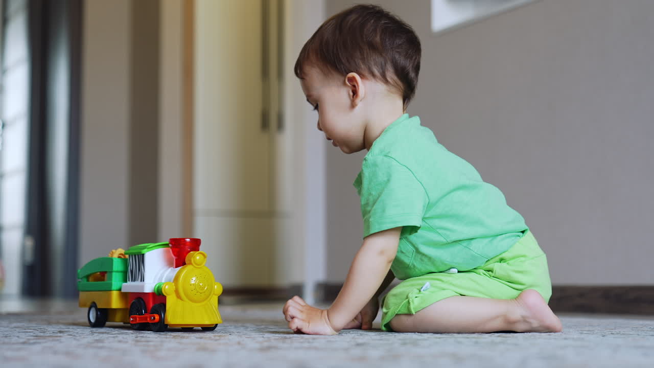 Cute baby boy presses button persistently on his toy. Adorable kid watches the toy train moves away and smiles. Low angle view.