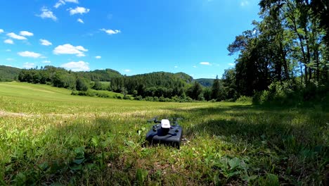 Un-Pequeño-Dron-Blanco-Con-Cámara-Está-Despegando-De-Una-Bolsa-Negra,-Un-Paisaje-De-Día-Soleado-En-El-Fondo-De-La-Naturaleza-Con-Bosques-Y-Prados-De-Hierba