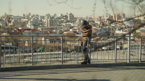 a father holds his son close to an iron railing as they both gaze out over a busy city, dressed warmly in jackets, with cars passing below with a tree branch behind