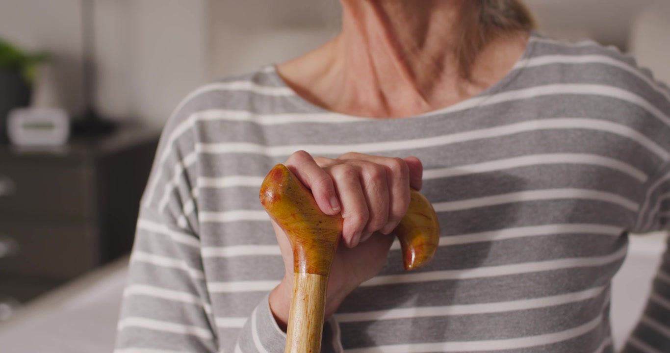 Una donna anziana tiene e stabilizza un bastone di legno, regolando la presa e la postura per l'equilibrio vicino al letto.