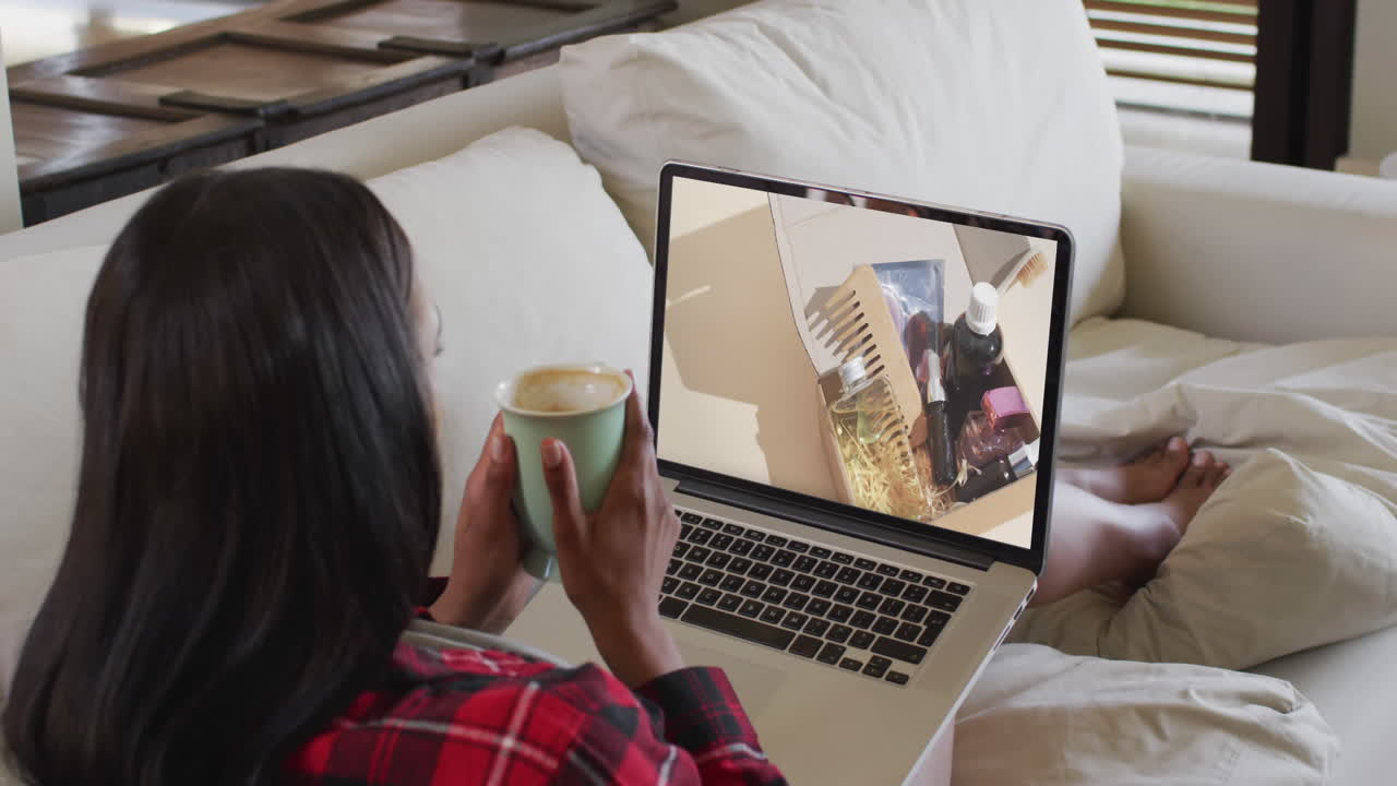 Biracial woman using laptop on couch at home online shopping for beauty products, slow motion