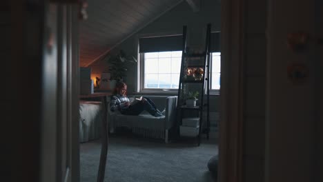 dolly shot of young girl laying on a small couch nex tto a window in a bedroom, reading a book, calm environment and surroudnings