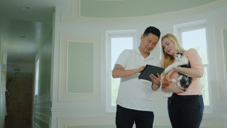 A-Family-Multi-Ethnic-Couple-Is-Holding-A-Cat-And-Is-Looking-At-The-Plan-Of-A-New-Home-On-A-Tablet-A