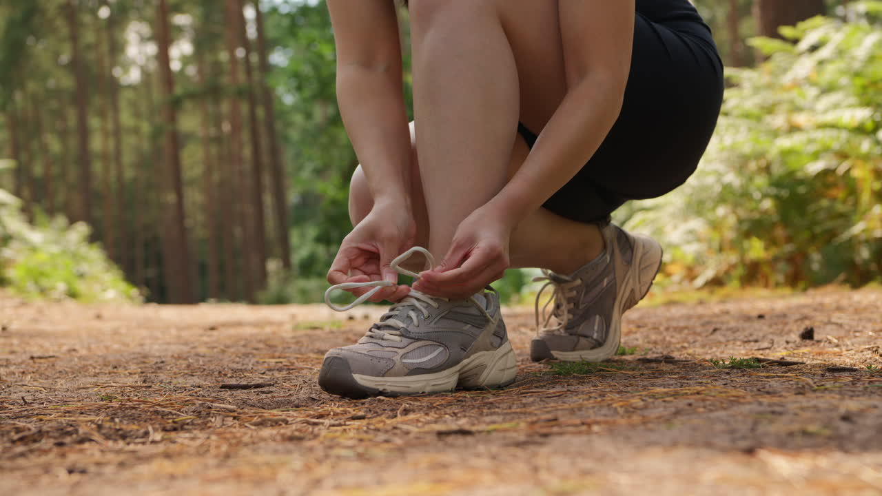 primer plano de una mujer atando los cordones de su zapato de entrenamiento antes de hacer ejercicio corriendo por la pista a través de un bosque filmado en tiempo real