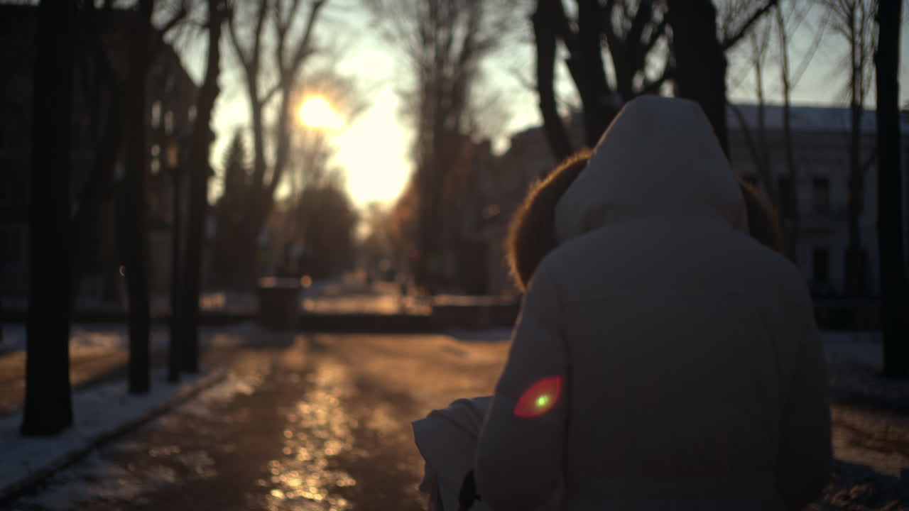 Rear view of a woman wearing a winter coat walking through a park at sunset, pushing a baby stroller