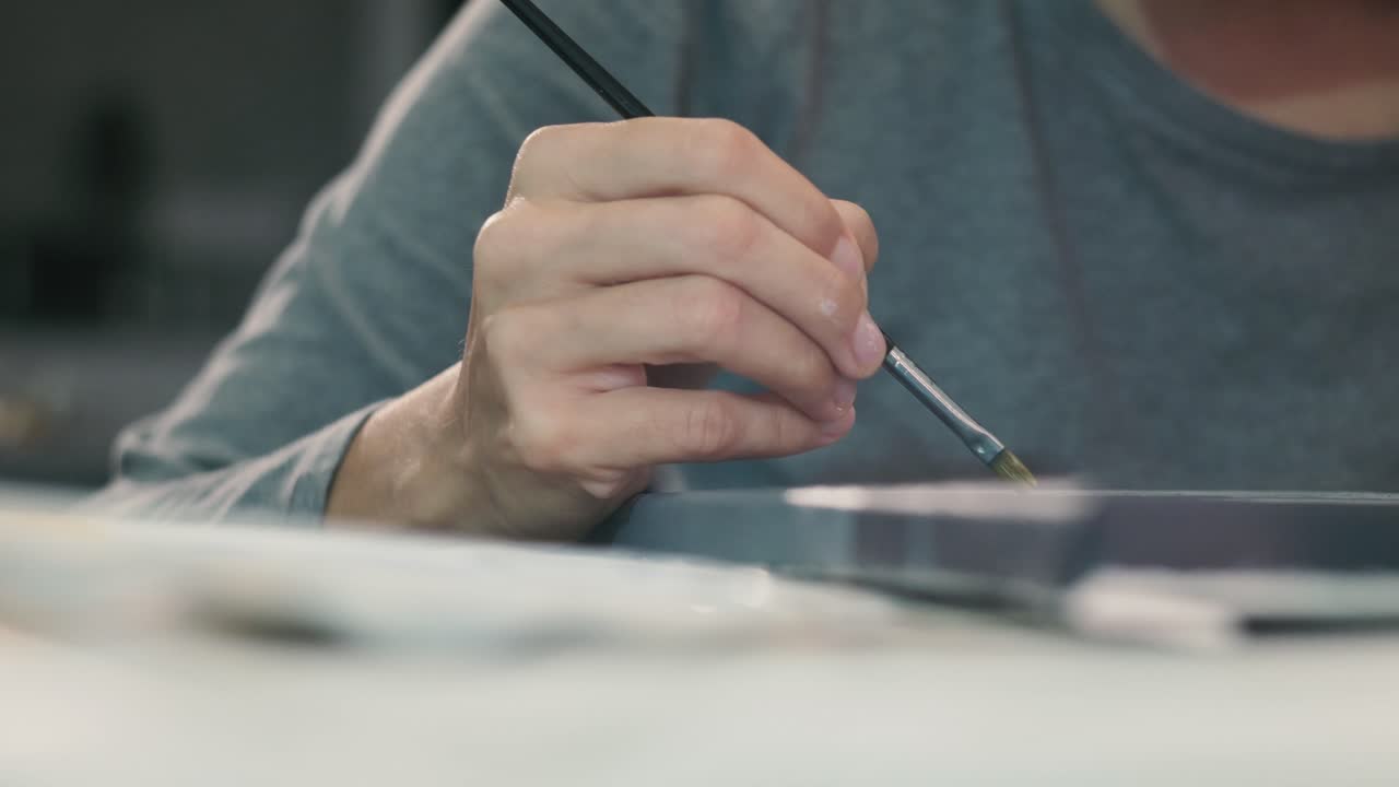 Young woman's hand holding a brush, drawing with paint from a palette in her art studio