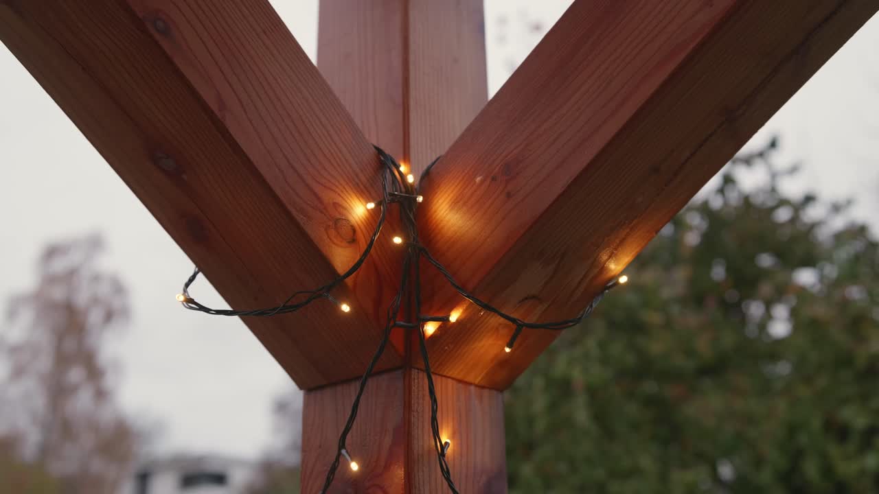 Christmas light chain being hung in the corner of a wooden pergola on a rainy, overcast November day. The lights are off at first, gently moved, then turn on as raindrops cling to the wooden supports