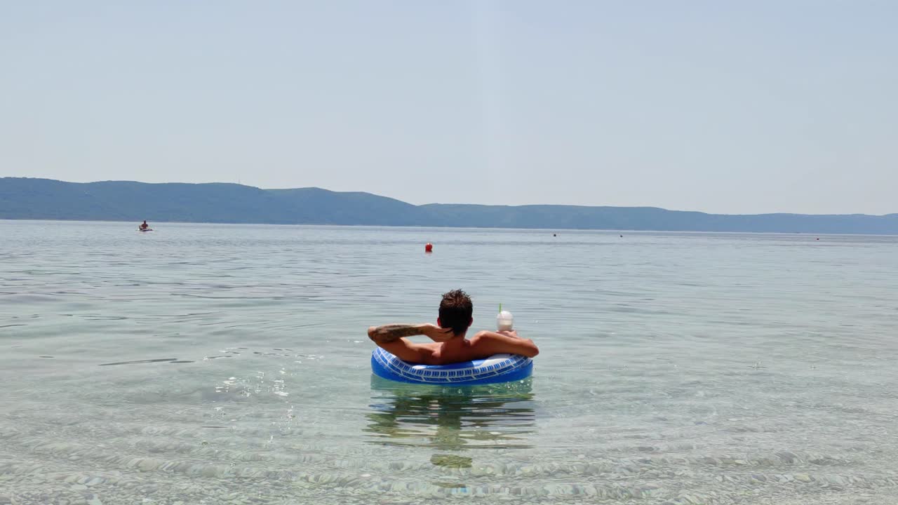 Static shot of guy on inflatable tube in the sea with drink relaxing in the sun