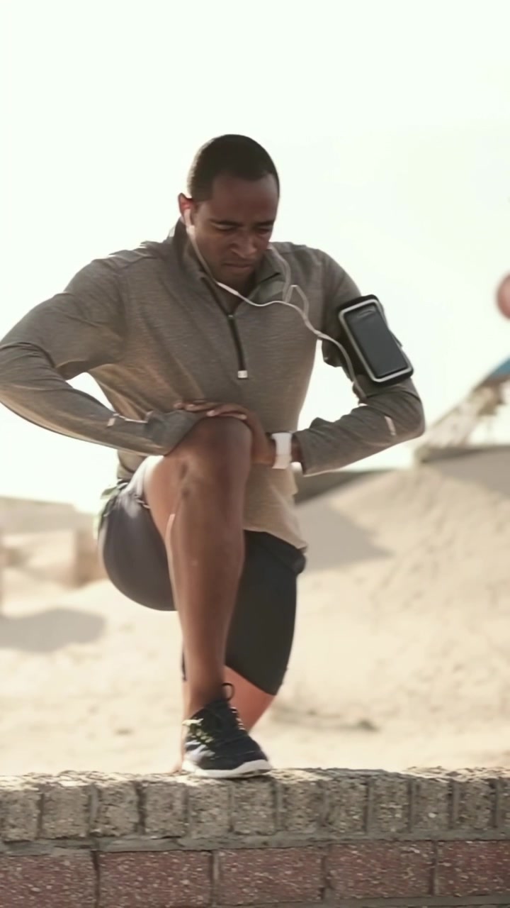Vertical video: Entering right, hooded runner warming knee stretch on wall at beach checking watch