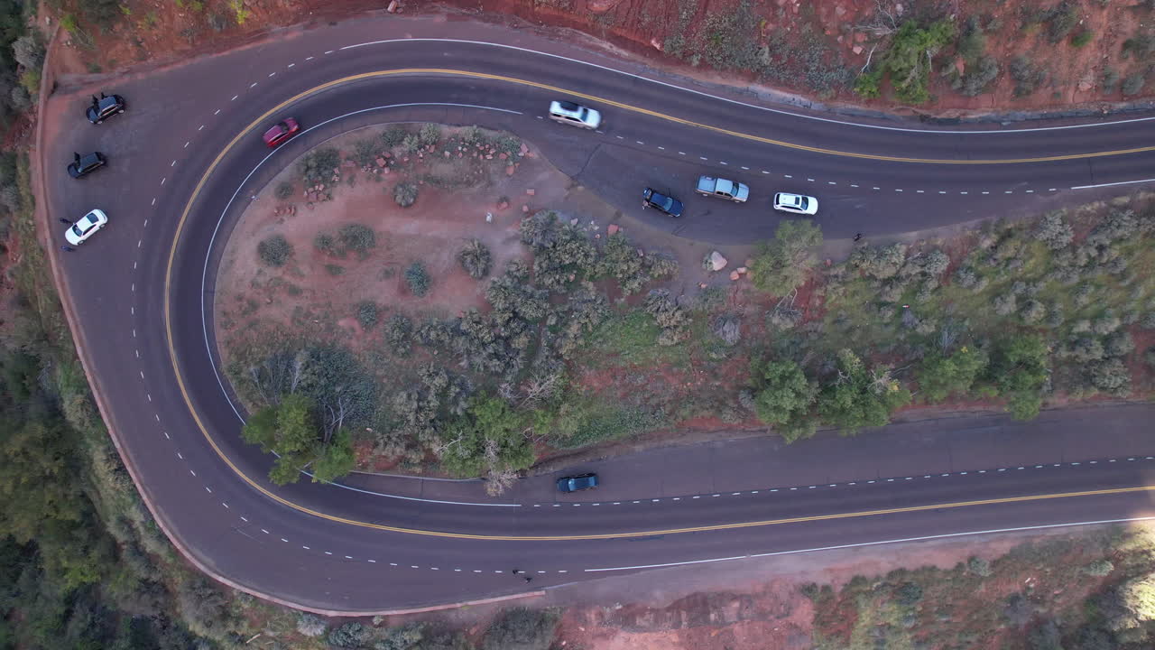 Overhead view of a curving mountain road with vehicles parked and driving, surrounded by desert vegetation and red rock terrain.