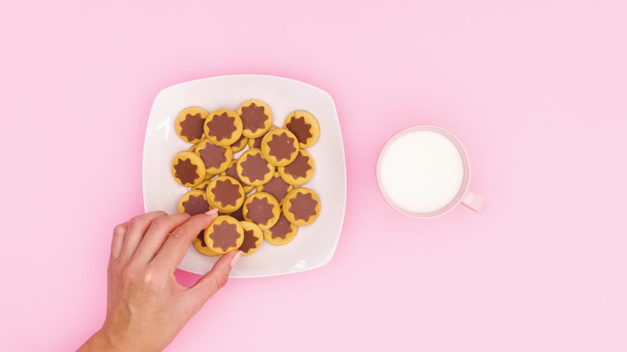 Chocolate cookies filling white plate and milk filing pink cup on pink theme and hand take one cookie and milk. Stop motion