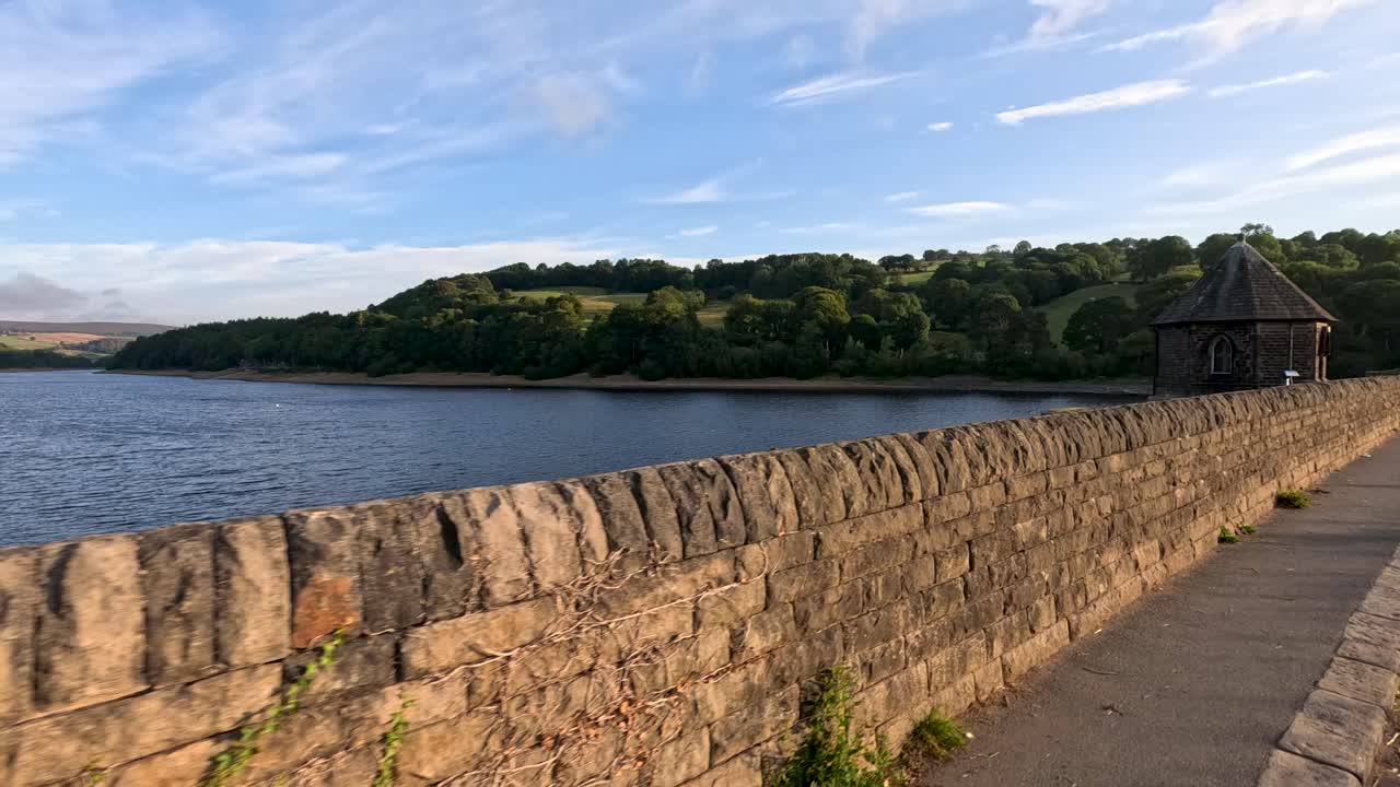 Camera moves smoothly along a stone bridge beside a river, with lush green hills and a blue sky in warm evening light