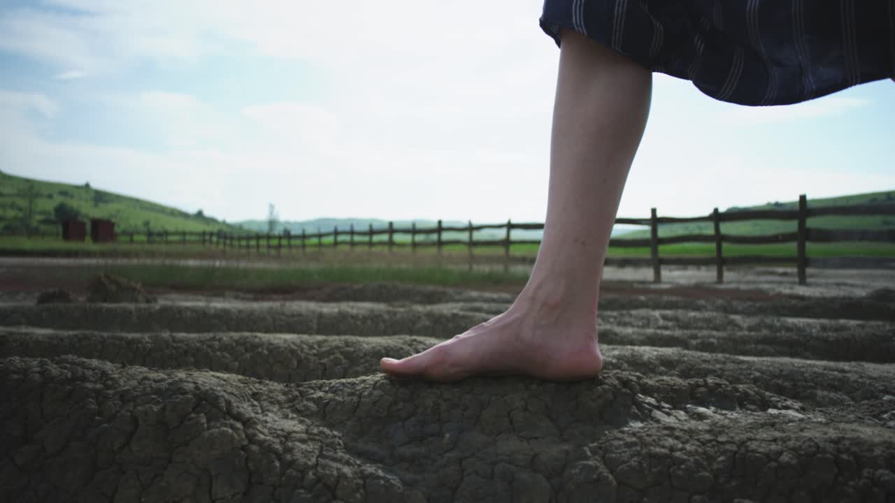 Low-angle close-up of a woman's bare feet carefully walking across the cracked, and dry terrain of a mud volcano. The scene evokes a sense of exploration and connection to a rare natural wonder.