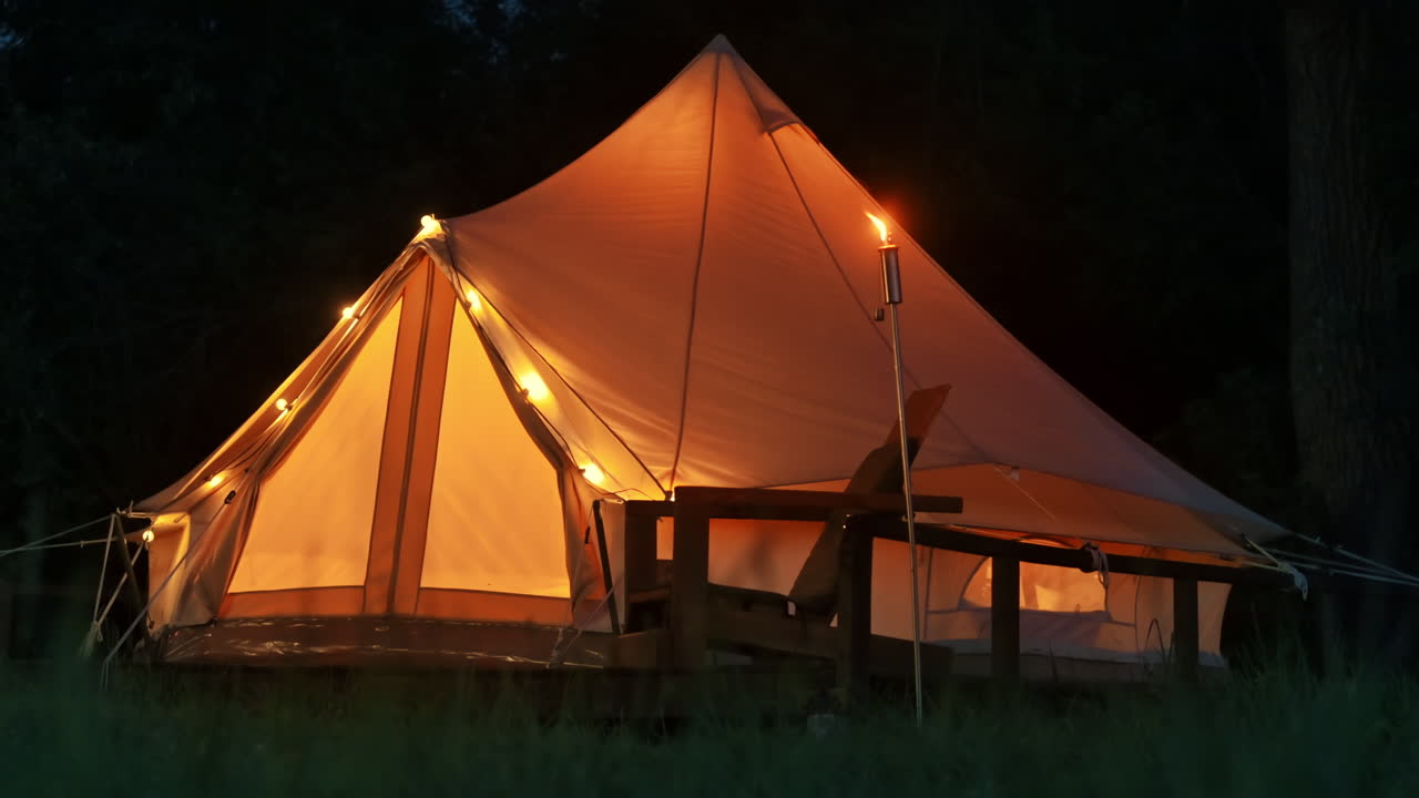 Telt med brændende fakler, lamper og træstole ved glamping, skov rundt om, nat