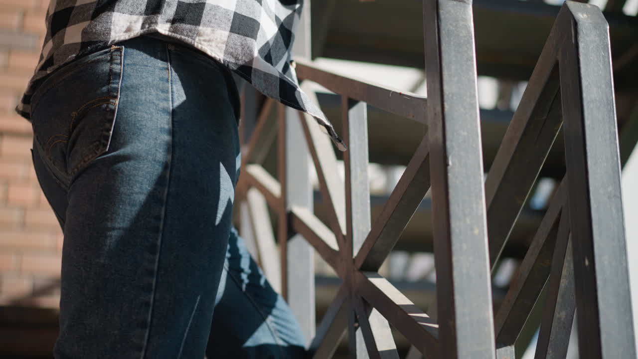 side view of man standing close to iron railing wearing blue jeans and black white plaid shirt with sunlight casting geometric shadows on brick wall urban stairwell