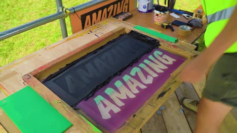 man wearing high vis painting printing font text signage words of positivity amazing manual labour with a piece of wood diy carpentry outdoor in nature scaffolding around tools next to him wristband