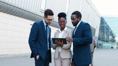 three multiethnic business people in stylish clothes reading something on the tablet and talking in the street