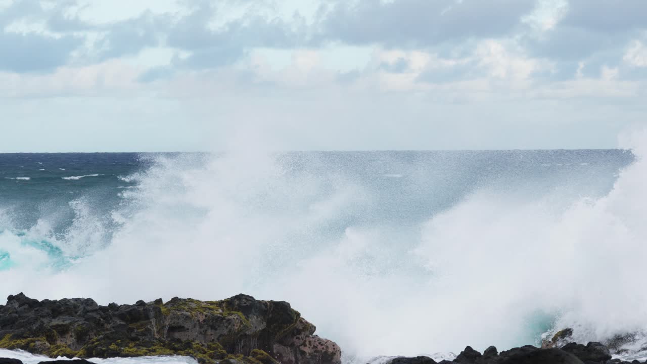 Powerful turquoise ocean waves crash forcefully against jagged black rocks, white surf spraying high across the rugged shoreline beneath a cloudy horizon