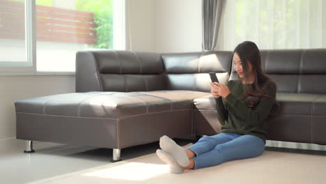 a pretty young woman sitting on the floor holding her smartphone laughs at what she sees