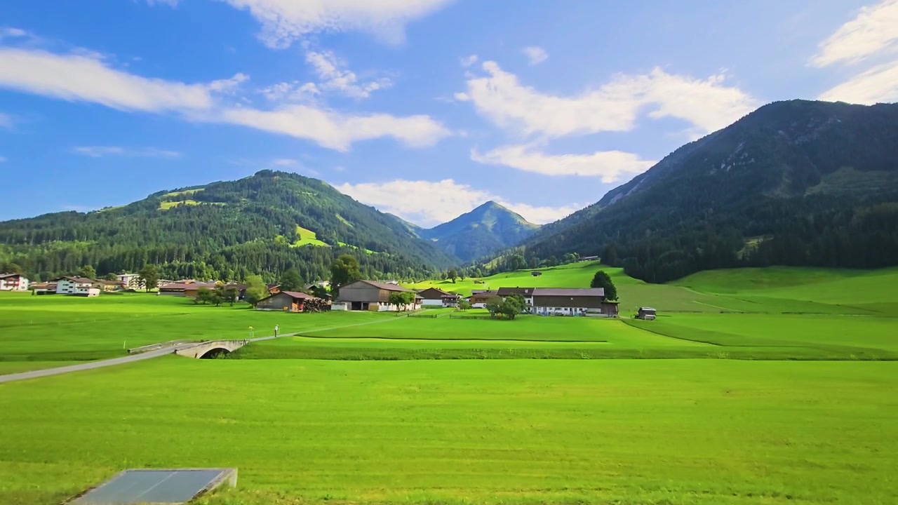 Viaggio in treno in Austria attraverso una tranquilla valle di montagna, che mostra prati rigogliosi, case in stile chalet, alberi a bordo strada e cime boscose sotto cieli estivi.