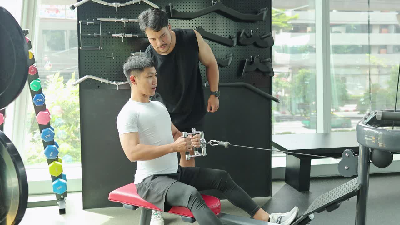 A man exercises on a rowing machine with a trainer's guidance in a well-lit gym environment