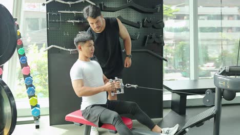 a man exercises on a rowing machine with a trainer's guidance in a well-lit gym environment