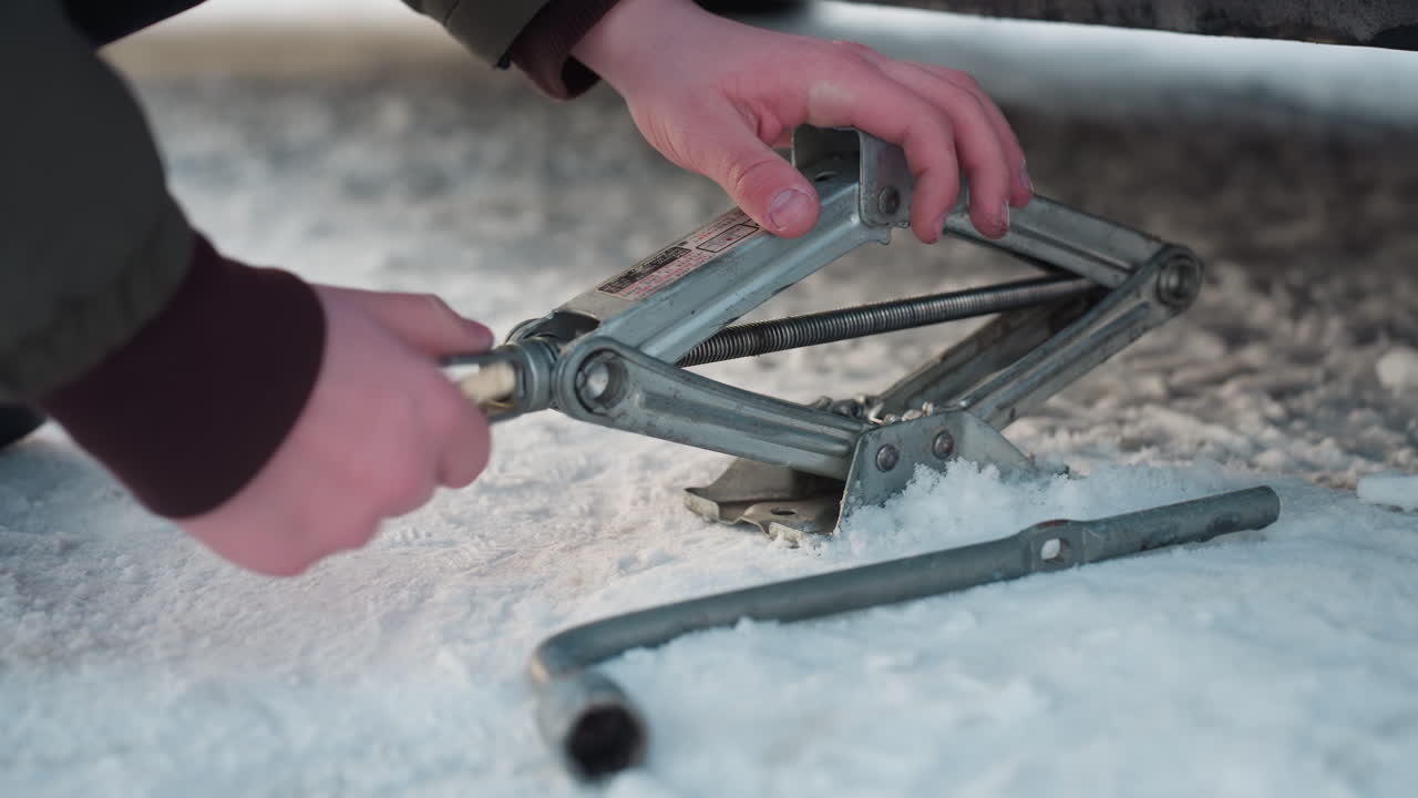 Close up of person adjusting jack on icy ground during vehicle maintenance, hands gripping metal parts while snow surrounds tool area, indicating preparation for lifting car in cold conditions