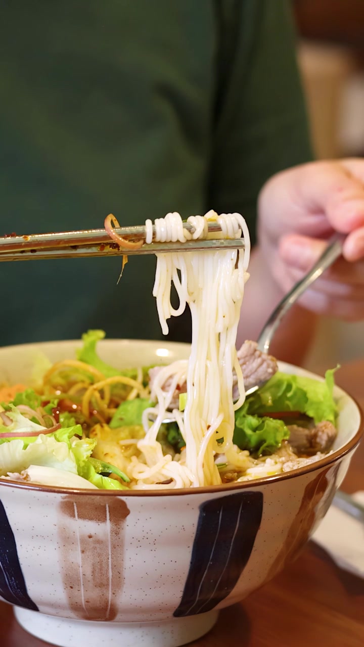 Primo piano di una persona asiatica che solleva spaghetti di riso da una ciotola di ceramica usando le bacchette