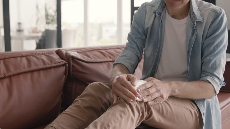 Uomo di mezza età che cerca di alleviare il dolore al ginocchio con un impacco di ghiaccio, seduto sul divano di casa.