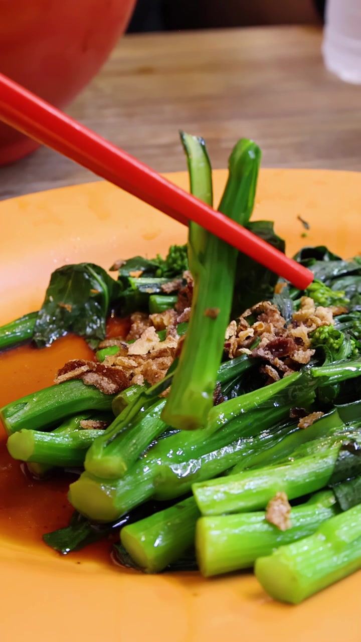 Bacchette rosse afferrano un pezzo di broccolo cinese da un piatto con salsa su un tavolo di legno, sotto un'illuminazione brillante e naturale con una bassa profondità di campo.