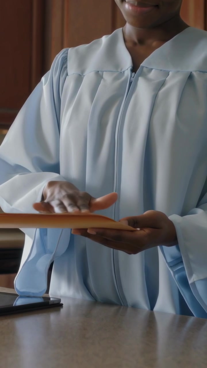 Vertical video: Holding manila envelope, graduate in gown smoothing it in kitchen, tablet nearby