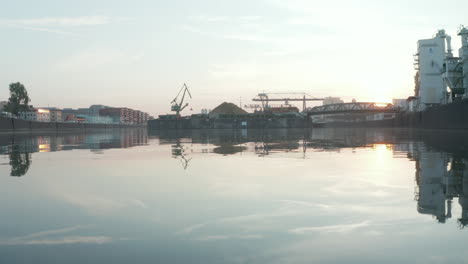 Una-Botella-De-Vidrio-Vacía-Flotando-En-El-Río-Con-Zona-Portuaria-Y-Contenedores-En-El-Fondo,-Bajo-Vuelo-Aéreo-Sobre-El-Agua-En-Los-Reflejos-De-La-Luz-Del-Sol-De-La-Mañana-Temprano