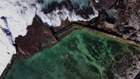 top down aerial of man swimming in tide pool on rocky coast with crashing waves