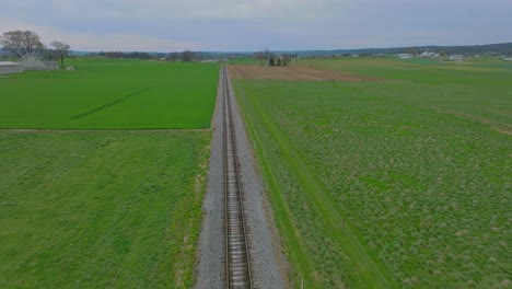 a drone view traveling along a single empty rail road track that goes thru green farmlands on a spring day